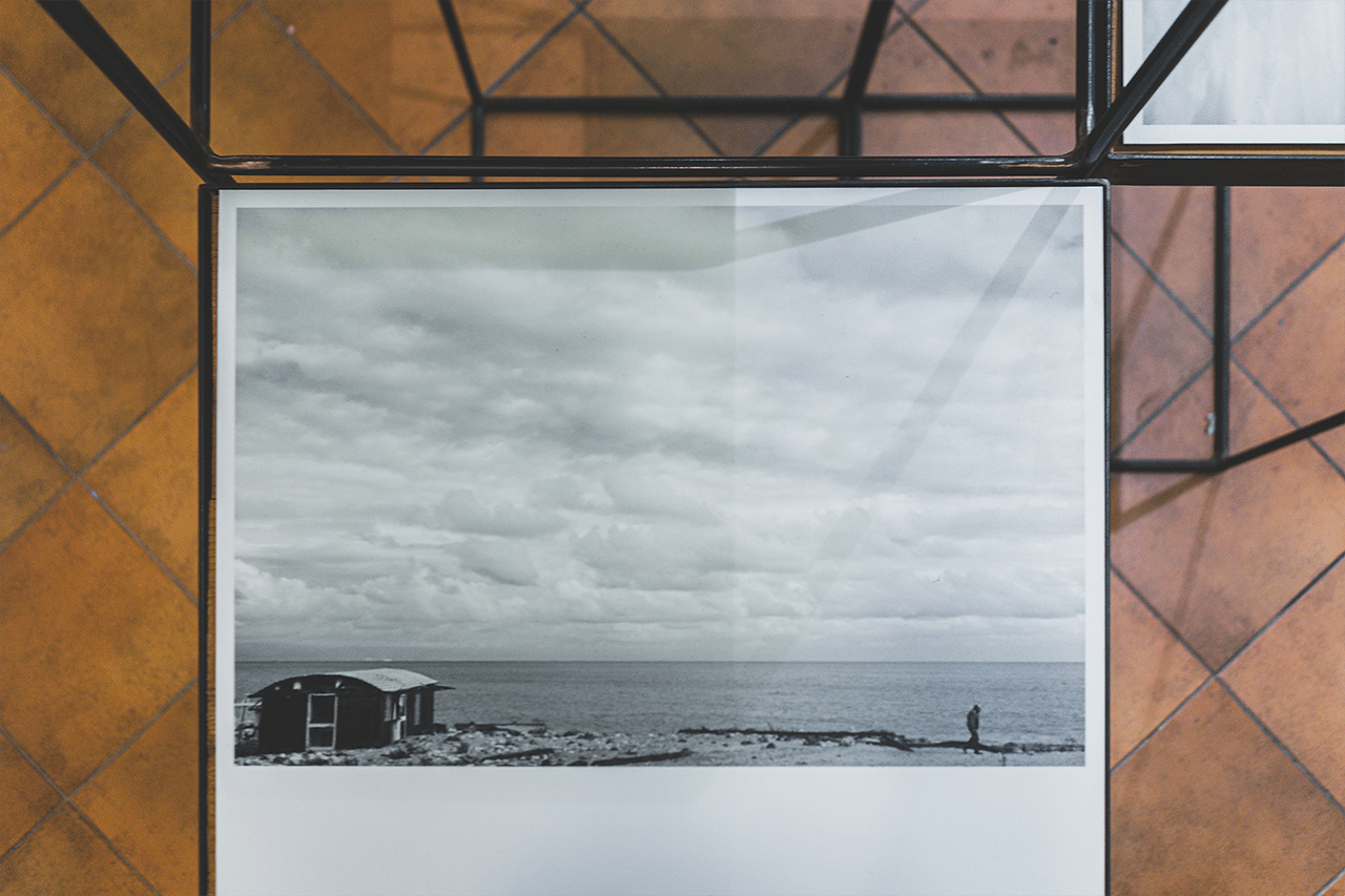 foto di marcello campora in esposizione che raffigura un piccolo capanno sul mare a sinistra, una persona che cammina a destra, un cielo pieno di nuvole
